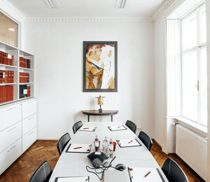 Gerade Ansicht auf das helle Besprechungszimmer der Rechtsanwaltskanzlei Unger mit länglichem Tisch, Schreibutensilien, einer Statue der Justitia, einem abstrakten Gemälde sowie einem Bücherregal.