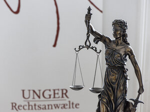 A photograph of a statue of Justitia, the goddess of justice and symbol of the legal system, in front of a roll-up banner bearing the law firm’s logo 